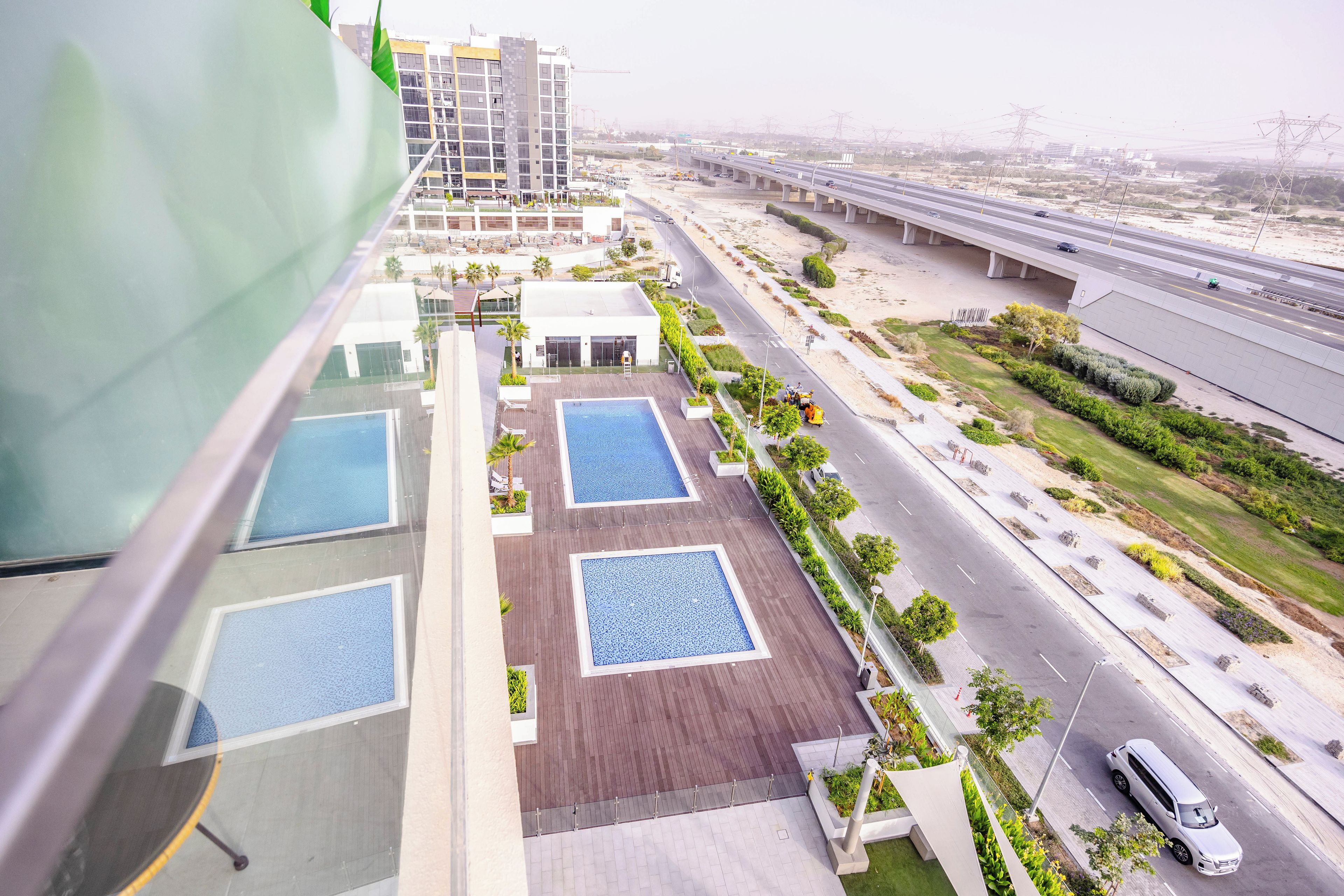 A high-rise building with multiple swimming pools, a landscaped area, and a busy highway in the background.