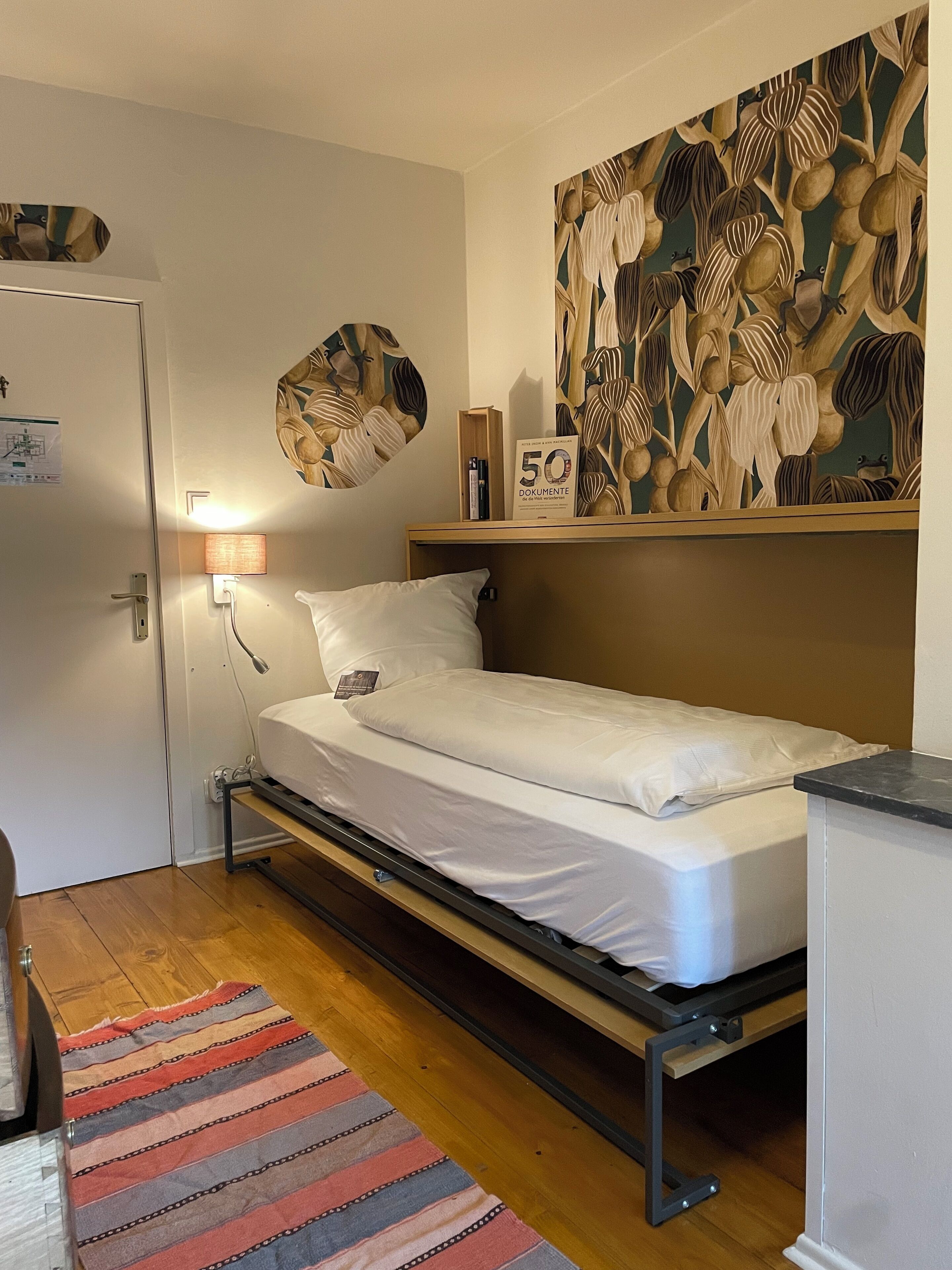 Un dormitorio pequeño y ordenado con una cama individual, una lámpara de noche, un estante en la pared con libros y un adorno decorativo.