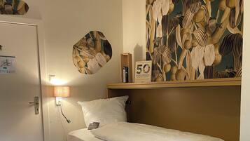 Un dormitorio pequeño y ordenado con una cama individual, una lámpara de noche, un estante en la pared con libros y un adorno decorativo.