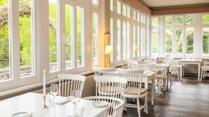 A dining area with white wooden tables and chairs, large windows, and candlelit lamps.