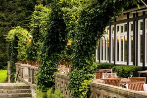 A garden with stone steps leading to wooden tables and chairs, surrounded by lush greenery and a building with large windows. - WaldesRuh am See (Aumuehle)