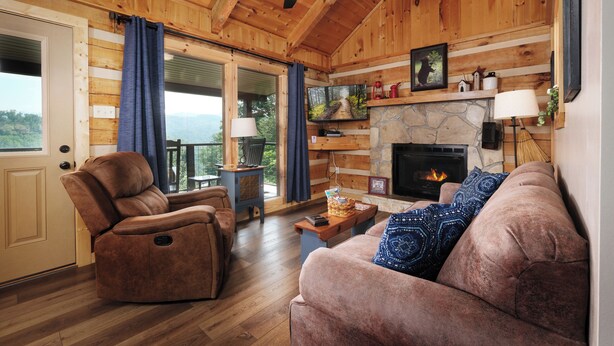 A cozy cabin interior with a fireplace, wooden walls, a recliner, a sofa, and a view through the door. - Clean and cozy one bedroom for 2- mountain view, hot tub, cute as can be! (Gatlinburg)