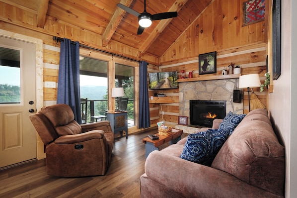 Interior acogedor de una cabaña con chimenea, paredes de madera, un sillón reclinable, un sofá y vistas a través de la puerta.