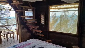 A rustic cabin room with a wooden staircase, a bed with a patterned bedspread, a wooden chair, and a large window offering a view of trees and a body of water. - Ecolodge Anjomba (Taolagnaro)