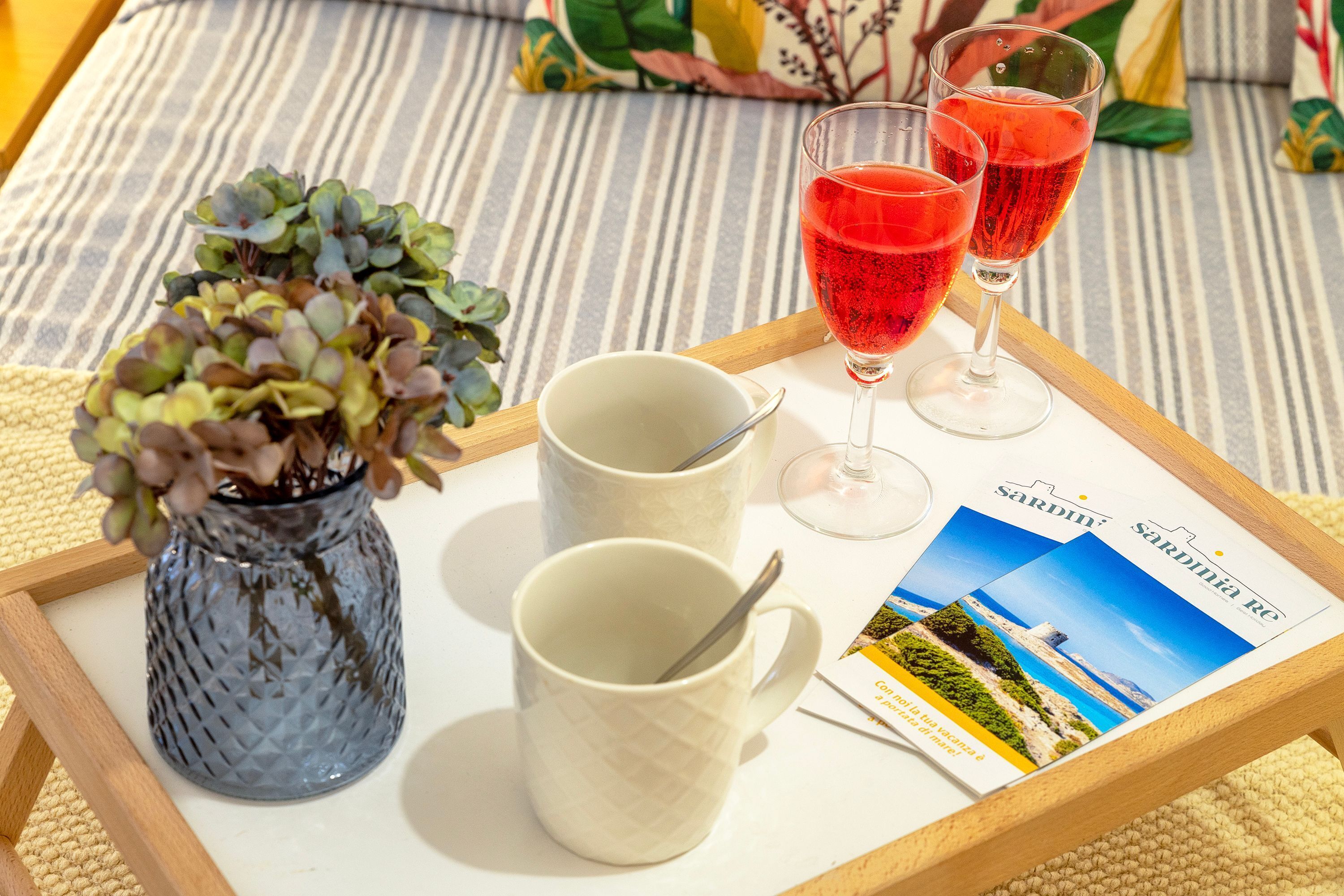 A tray with a vase of succulents, two white mugs, and two glasses of red drink on a striped sofa.