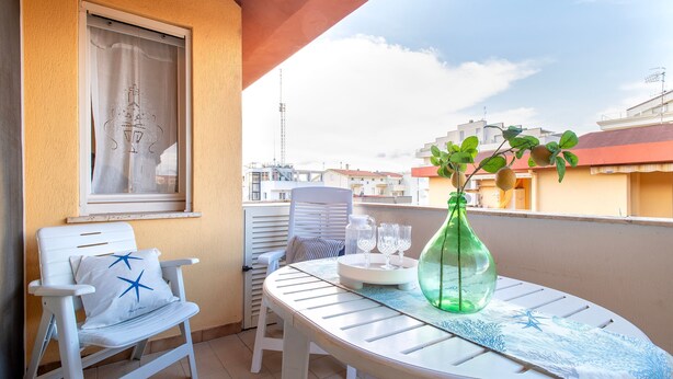 A balcony with a white chair, a table with a green bottle, and a window with a curtain. - Sardinia Re - Lido 68 (Alghero)