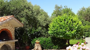 A stone-built outdoor oven with a tiled roof, surrounded by lush greenery and flowering plants. - Apartmani Luna (Sukosan)