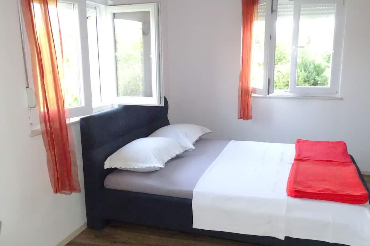 A single bed with white and red bedding, two white pillows, and a dark headboard in a room with white walls and wooden flooring.