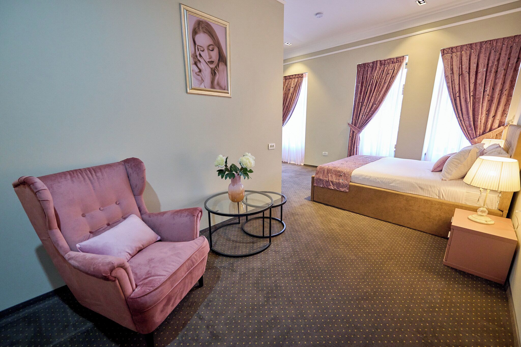 A hotel room with a pink armchair, a bed with a white comforter, a small table with a vase, and a nightstand with a lamp.