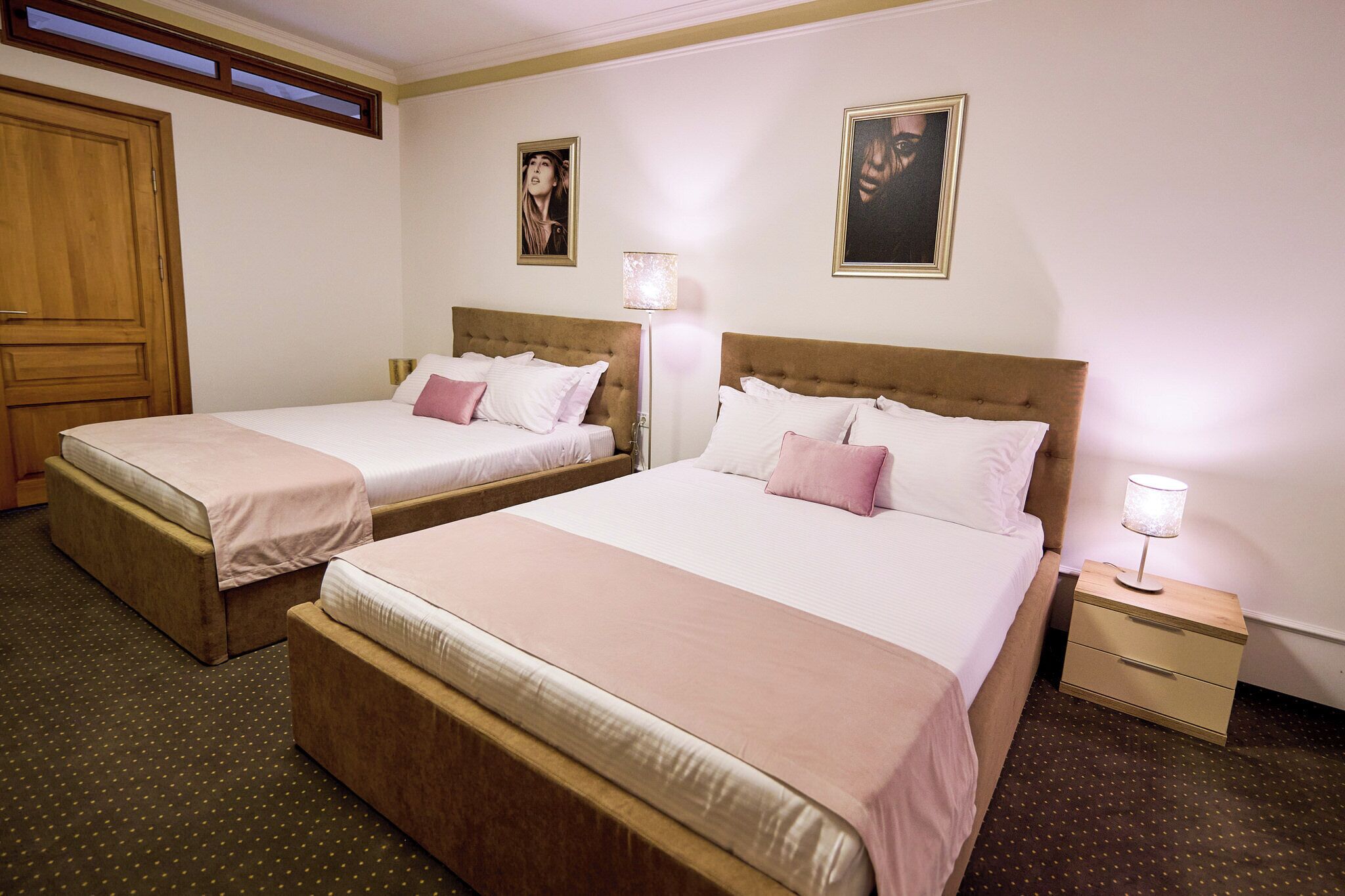 A hotel room with two beds, a wooden wardrobe, a bedside table with a lamp, and a framed picture on the wall.