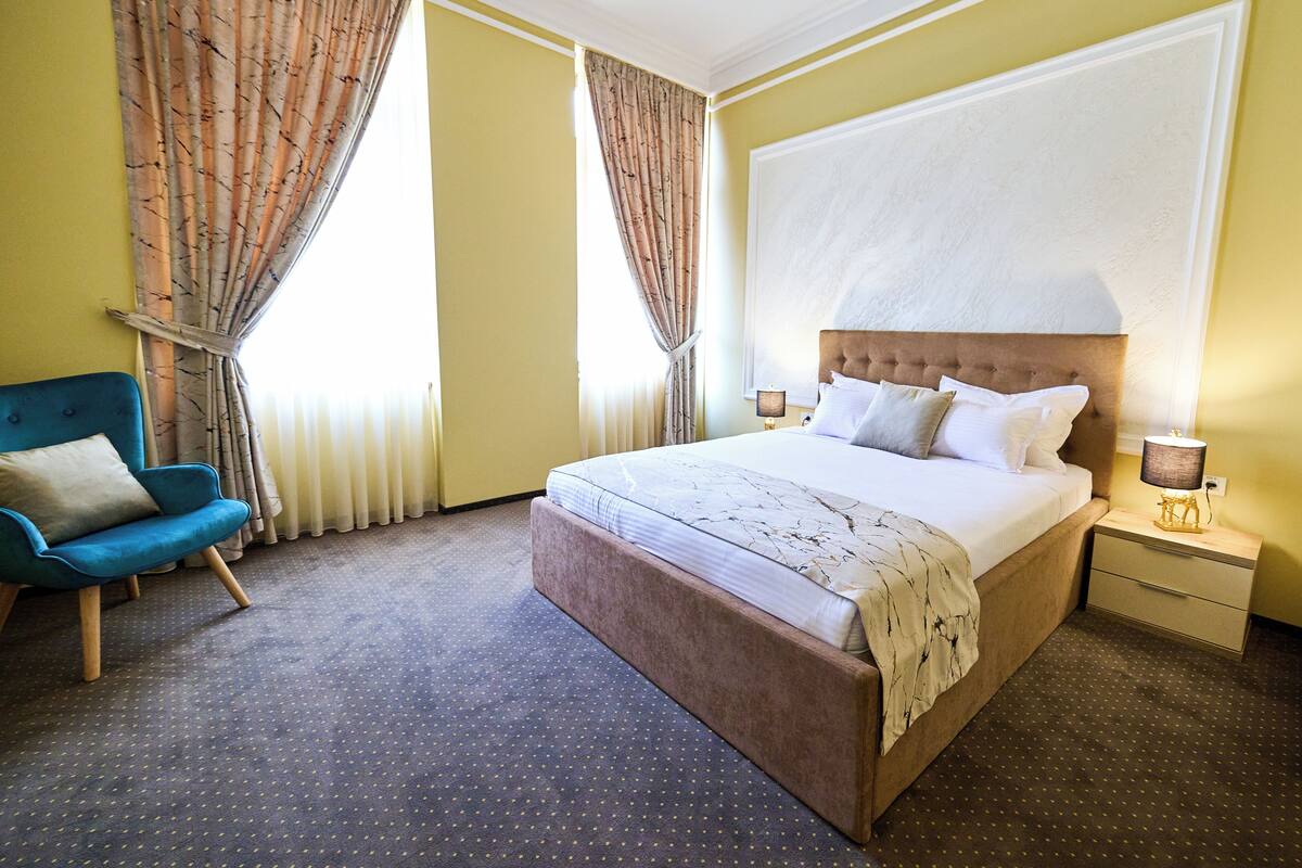 Une chambre d’hôtel avec un grand lit, un fauteuil bleu, une table de chevet avec une lampe et une fenêtre avec des rideaux.