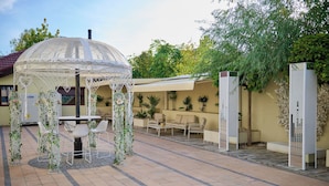 Een overdekt terras met een tafel en stoelen, omgeven door potplanten en een decoratieve gazebo.