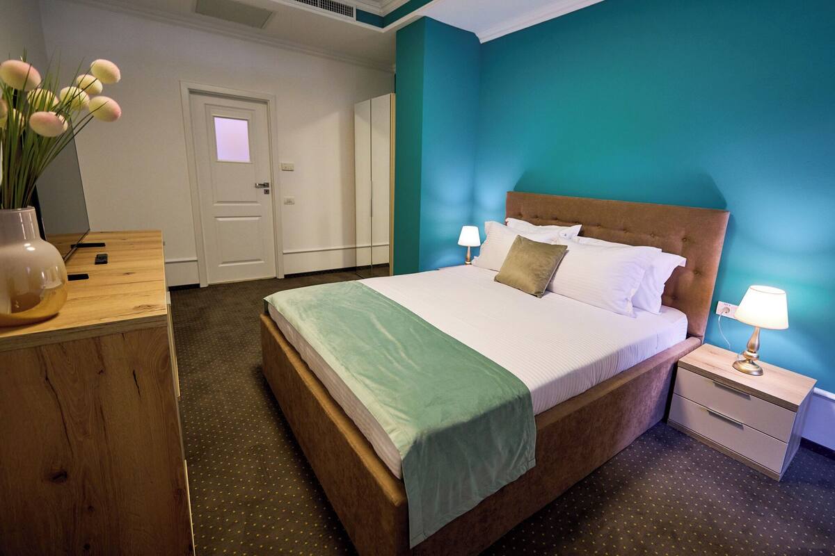 Une chambre d’hôtel avec un lit, des tables de chevet, une commode en bois, un vase avec des fleurs et des murs de couleur turquoise.