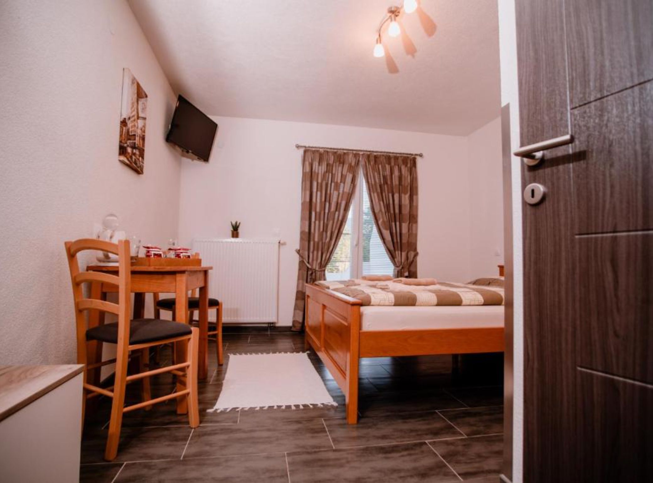 Habitació petita i acollidora amb un llit, un escriptori i una cadira. Hi ha una finestra amb cortines, un radiador i una televisió penjada a la paret.