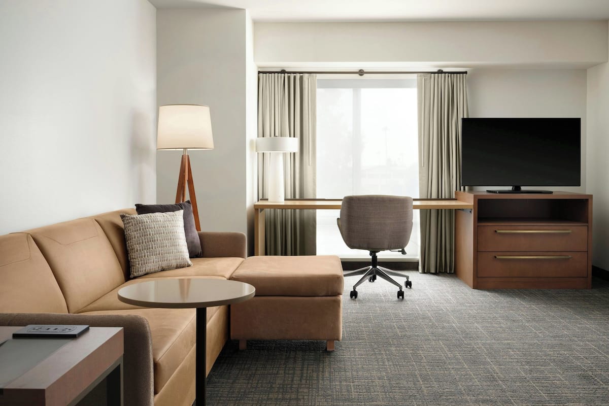 Una habitación de hotel con sofá, reposapiés, escritorio, silla, televisor, lámpara y una mesita.