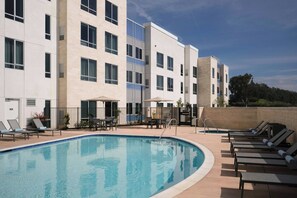 Una piscina con sillas de descanso y sombrillas, rodeada por un edificio con varias ventanas y balcones.