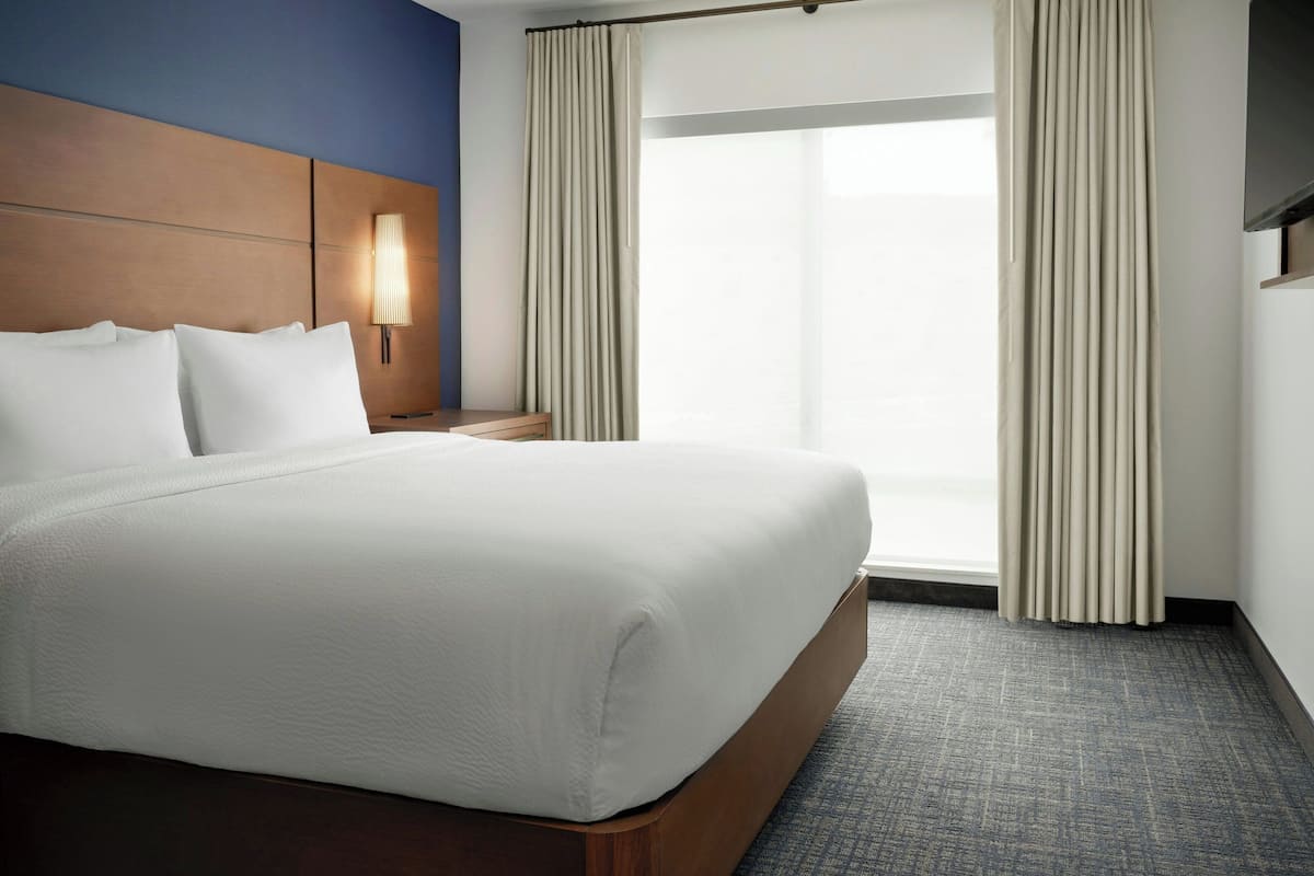 Habitación de hotel con una cama grande, una mesita de noche, una lámpara de pared y un ventanal con cortinas.