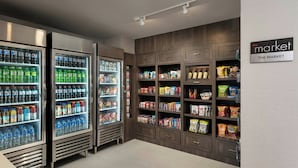 Interior de una tienda de comestibles con dispensadores refrigerados de bebidas, estantes con bocadillos y un cartel que dice 'the market'.