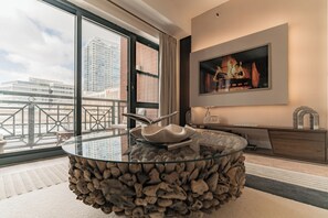 Ein modernes Wohnzimmer mit einem Glastisch, der von Steinen umgeben ist, einem an der Wand befestigten Flachbildfernseher und einem Balkon mit Stadtblick.