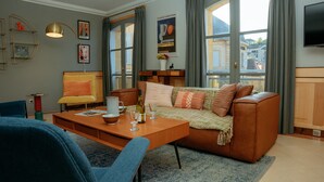 Un salon avec un canapé en cuir marron, une table basse en bois et un fauteuil bleu. Deux tableaux encadrés ornent le mur, ainsi qu’un vase rempli de fleurs et une fenêtre avec des rideaux.