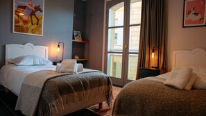Hotellihuone, jossa on kaksi sänkyä, suuri ikkuna, yöpöytä ja taideteoksilla koristeltu seinä.