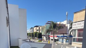 A balcony with two white plastic chairs, a green artificial turf floor, and a view of a street with buildings and shops. - Balcony Studio Vacation Home (Unknown)