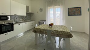 A modern kitchen with white cabinets, a microwave, and an oven. There is a dining table with a patterned tablecloth and chairs. A clock and a window with curtains are visible on the wall. - Pearl Diva Apartment (Palermo)