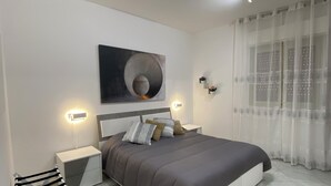 A modern bedroom with a large bed, bedside tables, a wall-mounted abstract art piece, and sheer curtains. - Pearl Diva Apartment (Palermo)