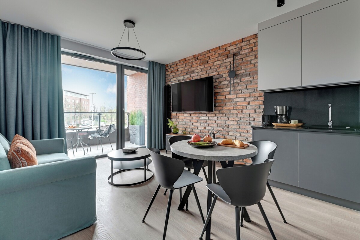 A modern living space with a dining area, a kitchen with a brick wall, and a large window offering a view of the outside.