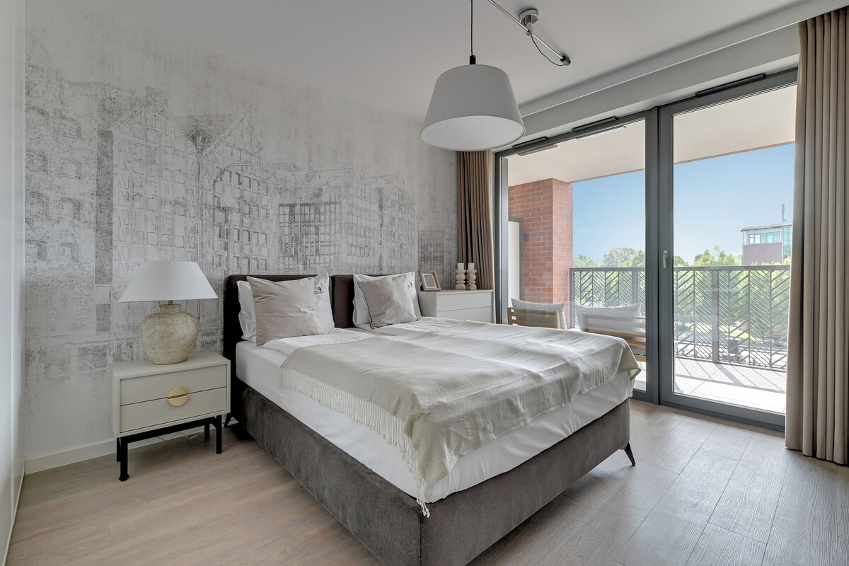 A bedroom with a large bed, a nightstand, a lamp, and a view of a balcony.