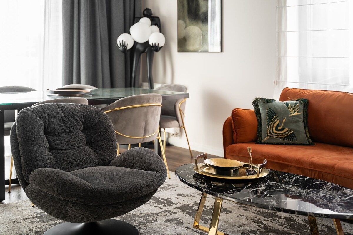 A modern living room with a dark grey armchair, a round table with a marble top, and an orange sofa with a decorative pillow.