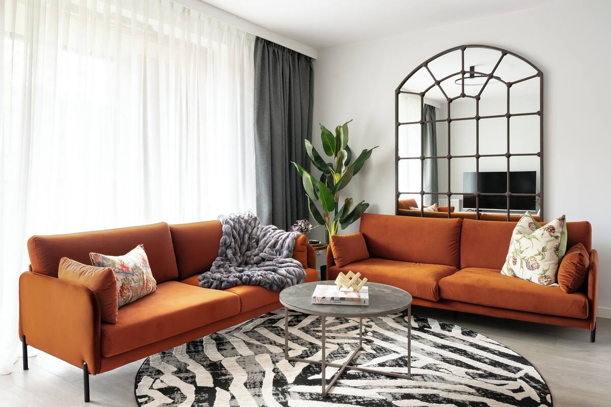 Ein Wohnzimmer mit zwei orangenen Sofas, einem runden Couchtisch, einer großen Pflanze und einem schwarz-weißen Teppich.