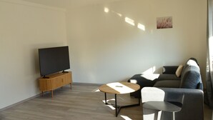 A modern living room with a flat-screen TV on a wooden stand, a grey sofa, a small round coffee table, and a large window allowing natural light. - Vacation apartment Taubersonne (Igersheim)