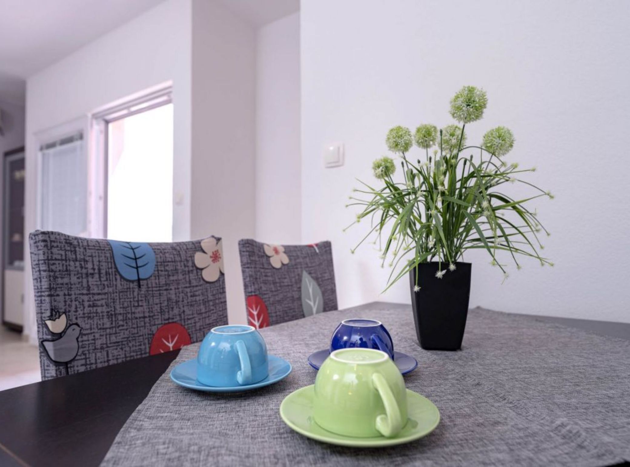 A dining table with a potted plant, three cups, and two patterned cushions.