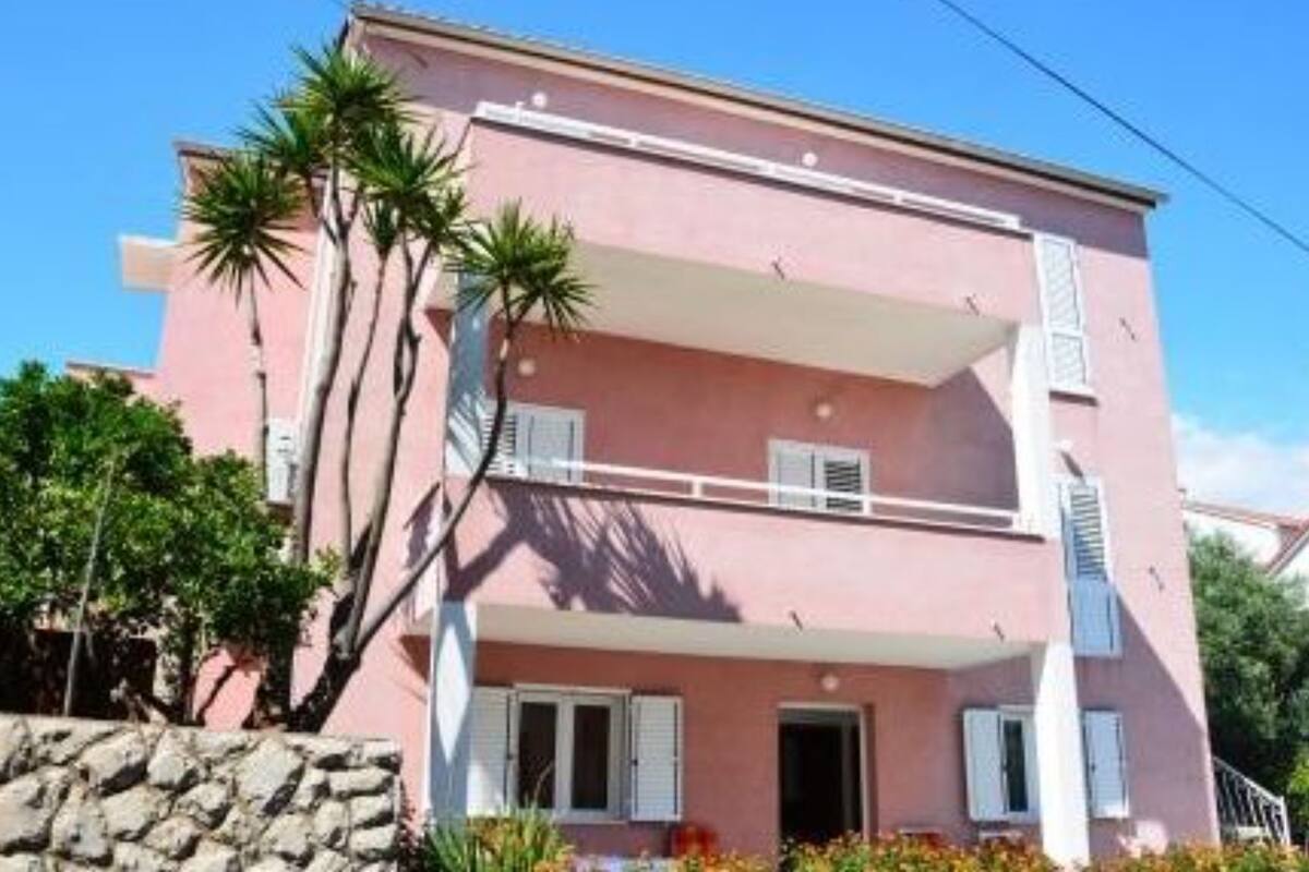 A two-story pink building with white shutters, a balcony, and a stone wall in the foreground.