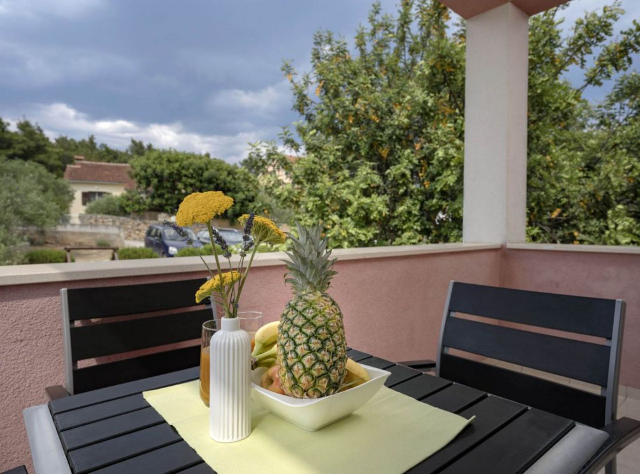 A table with a bowl of fruit and a vase of flowers on a balcony with a view of trees and a building.
