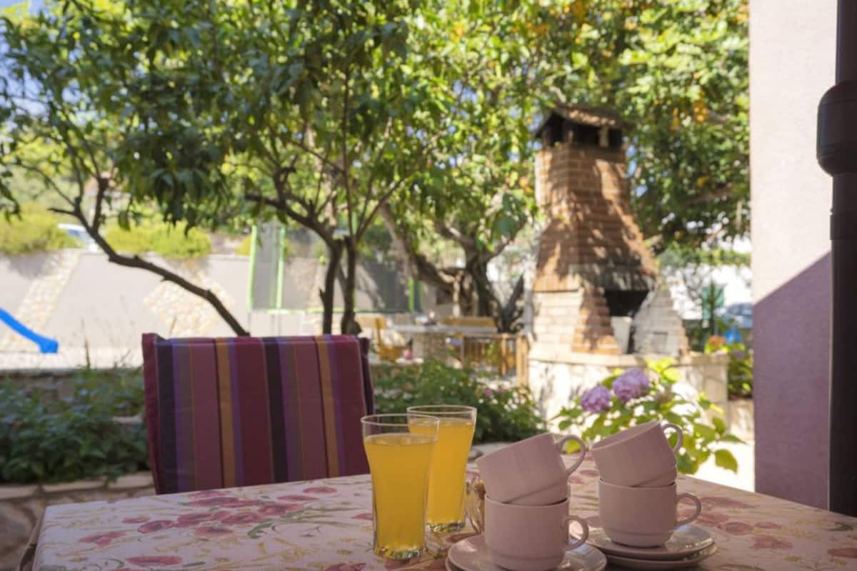 A table with a floral tablecloth, two glasses of yellow liquid, and three stacked cups on a patio with a view of trees and a brick structure in the background.
