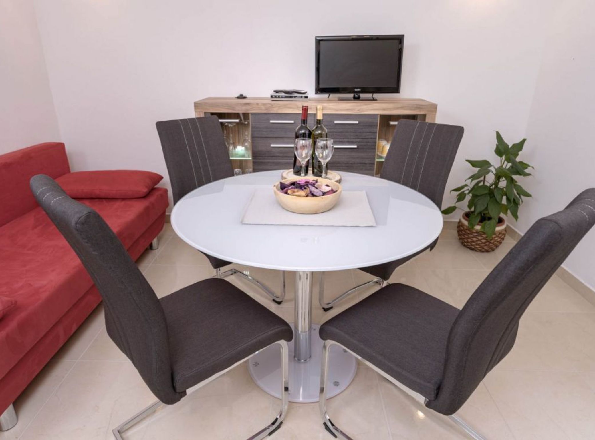 A modern dining area with a round table, four chairs, a red sofa, a TV, and a plant.