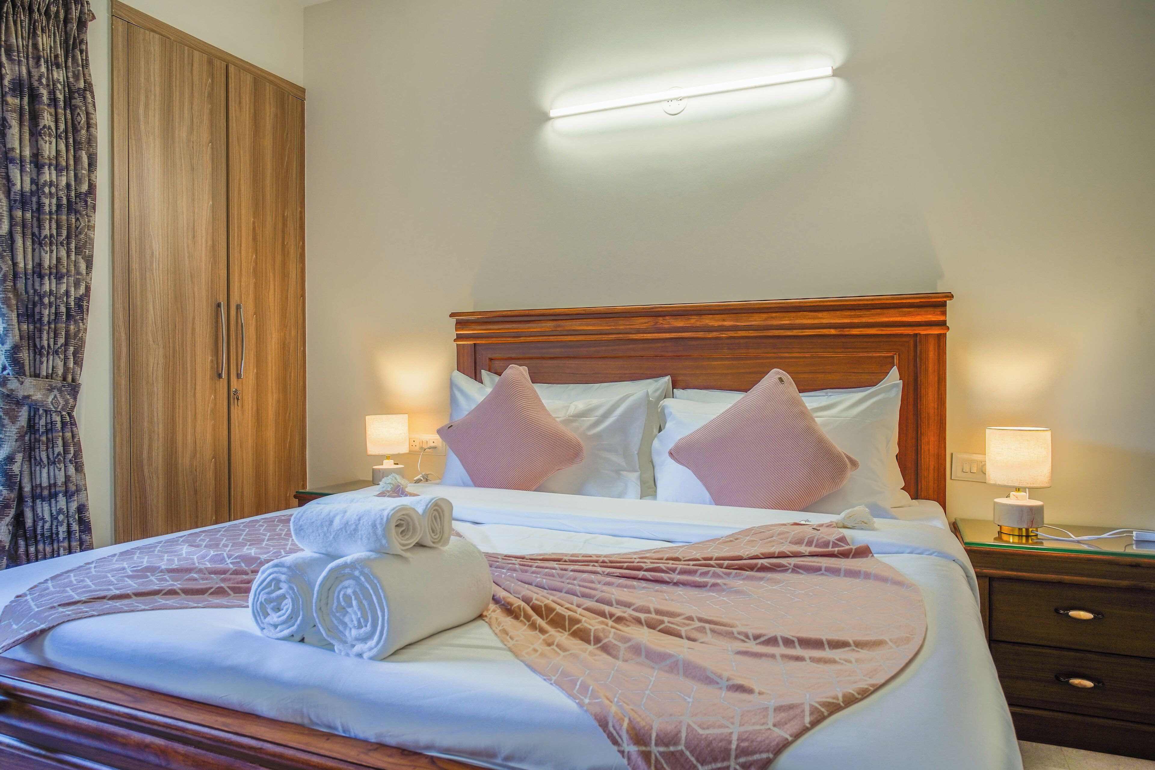 A neatly made bed with rolled towels, a wooden headboard, and bedside lamps.