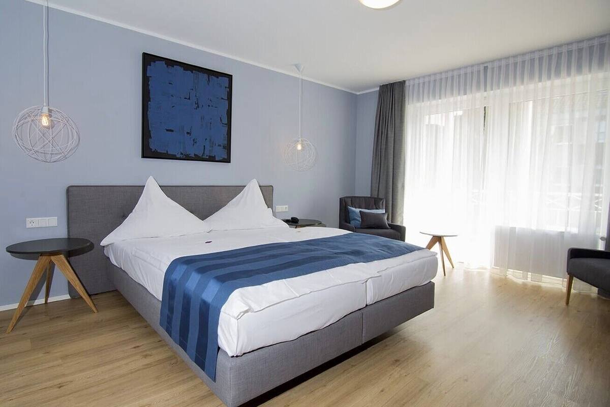 Un dormitorio moderno con una cama grande, dos mesitas de noche, una silla y un ventanal con cortinas traslúcidas.