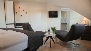 A modern hotel room with a bed, two chairs, a small table with a bottle and glass, a TV, and a kitchenette. - Hotel Fliederbusch  (Borken)