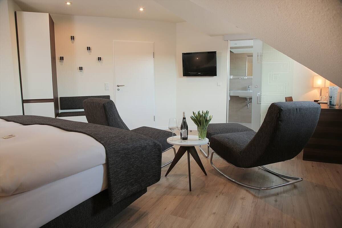 Habitación de hotel moderna con una cama, dos sillas, una mesita con una botella y un vaso, televisor y kitchenette.