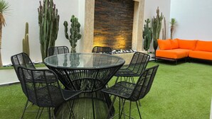Un espai interior modern amb una taula de vidre envoltada de cadires, una paret de maó amb un relleu i diversos cactus.