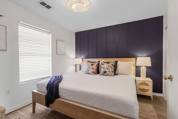 Quarto com cama, mesas de cabeceira, janela com persianas e painel de parede escuro.