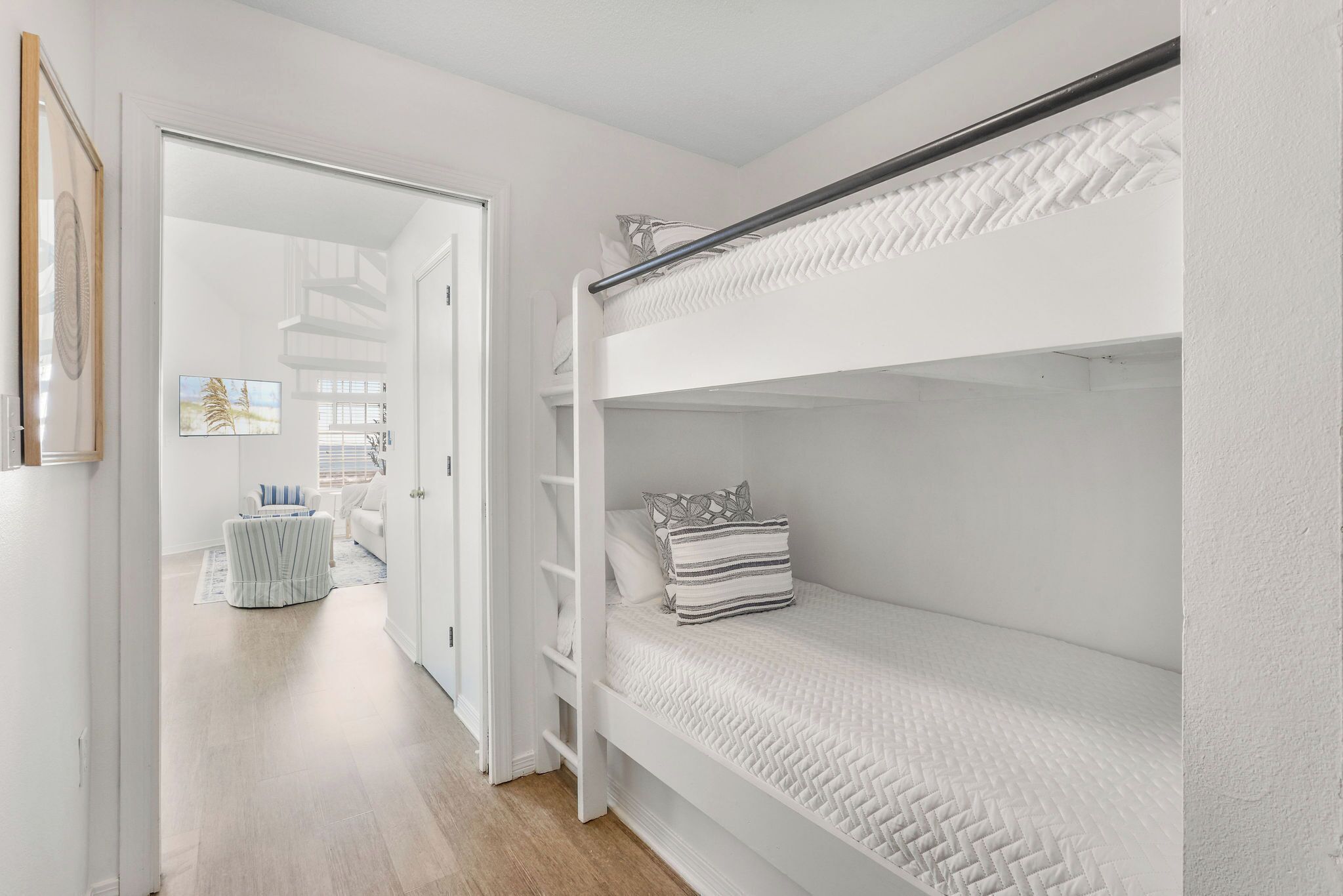 Una habitación blanca con literas, una puerta que da a otra habitación y una pequeña mesa con una silla.