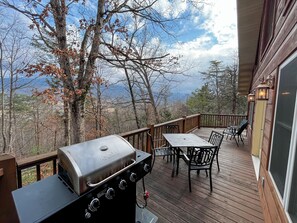 A deck with a grill, outdoor table and chairs, and a view of trees and mountains.