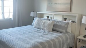 Un dormitorio ordenado con una cama grande, un ventilador de techo, una ventana con cortinas y un cuadro colgado en la pared.