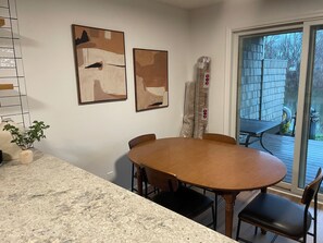 Een eethoek met een ronde houten tafel en stoelen, twee abstracte schilderijen aan de muur, een potplant en uitzicht door een glazen deur.