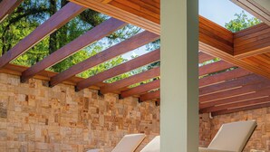 Piscina con acqua cristallina, pergolato in legno, muro in pietra e poltroncine da relax bianche.