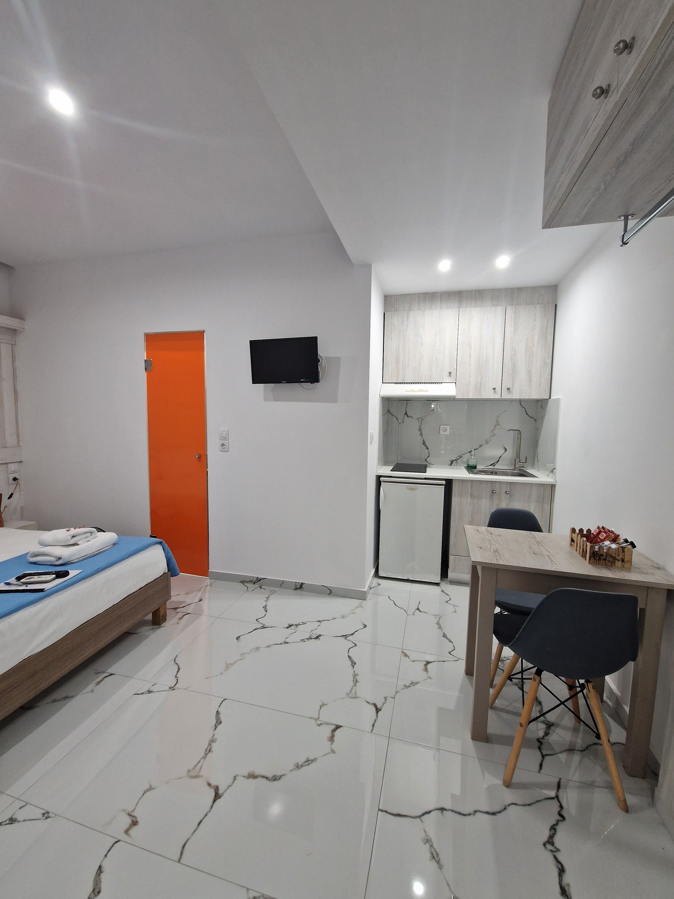 A compact hotel room with a bed, a small kitchenette, a dining area, and a TV mounted on the wall.